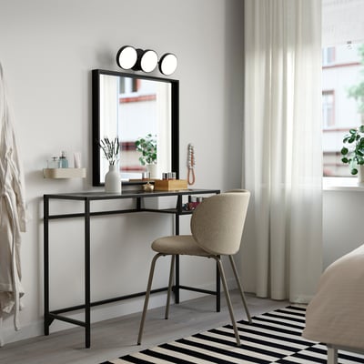 Black VITTSJÖ desk with glass top, chair, and mirror with lights, near large window.