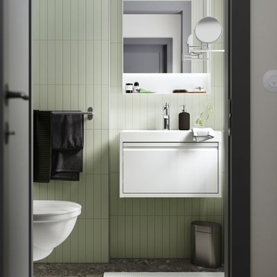 The modern bathroom a white ÄNGSJÖN vanity with sleek cabinetry, smooth basin, neatly hung black towel, metallic accents, and minimalist green tiles.