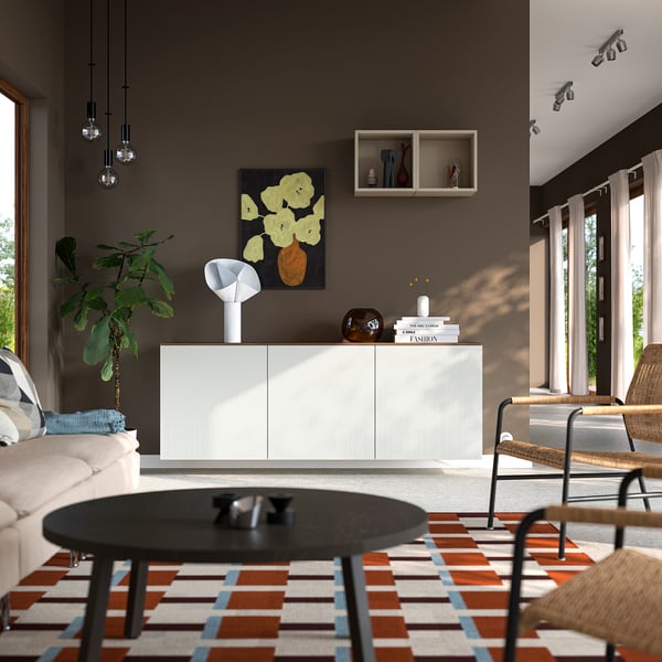 Modern living room with BESTÅ sideboard, books, black coffee table, beige sofa, orange rug, wicker chair, dark walls, light curtains, art.