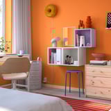 Orange room with white EKET shelves holding books, vases, and decor. Blue stool and wooden chair. Yellow wall light above.