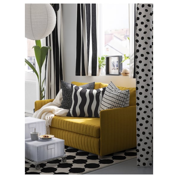 Modern living room with yellow sofa, black and white patterned curtains, pillows, and rug. Large windows let in natural light.