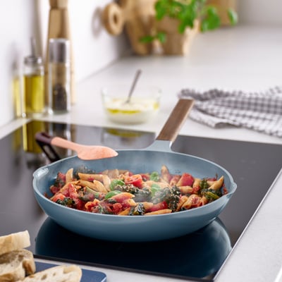 Blue cast aluminium frying pan with wooden handles, non-stick coating, on a kitchen counter, surrounded by food and kitchenware.