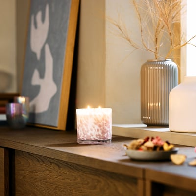 A cosy KRONSÅNGARE pink floral scented candle on a wooden table with fruit bowl and tall vase.