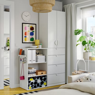 Modern bedroom with white furniture. Wardrobe, drawers, bookshelf, and plant by the window.
