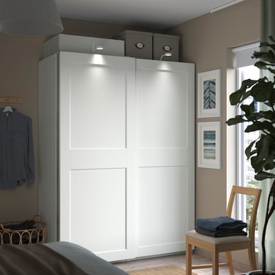 A modern bedroom with a white PAX wardrobe featuring sliding doors and built-in lights, flanked by decorative items and a window.