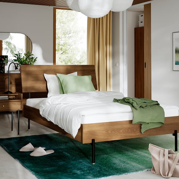 Bedroom with RÅDMANSÖ bed, green throw, white linens, wooden headboard & black legs. Rug & slippers nearby. Yellow curtains & floor lamp visible.