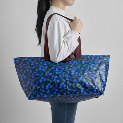 Person holding large, foldable SPJUTSÖ bag. Bag pattern is blue spots on dark background. Bag is easy to clean and store.