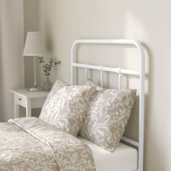 A bed with a white metal frame, beige patterned bedding, and pillows. A white lamp and small vase with flowers on a bedside table.
