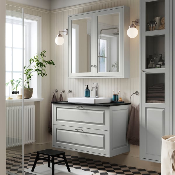 Modern bathroom with light grey TÄNNFORSEN vanity, sink, mirror, & storage. Wall-mounted vanity, black stool, white radiator, plants.