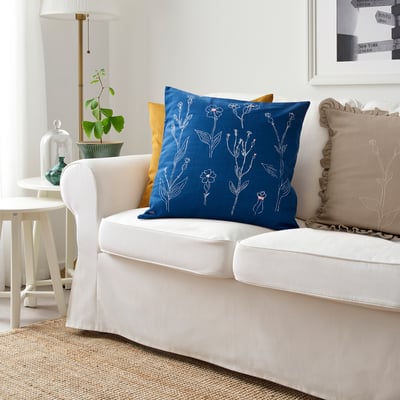 Modern living room: white sofa with blue floral cushion, beige pillow, mustard pillow. IKEA ÅKERNEJLIKA in 20x20 size, embroidered flowers.