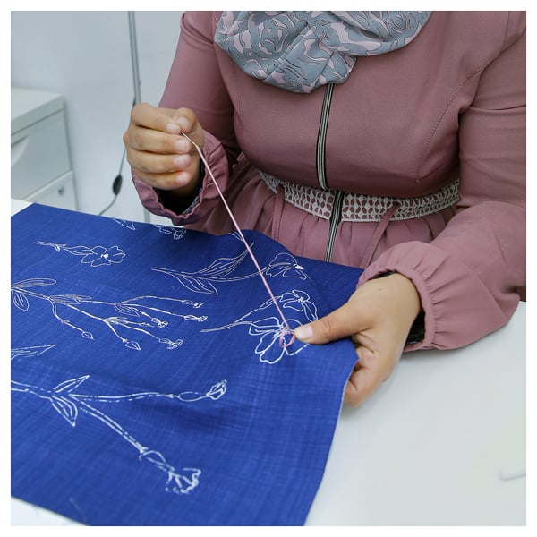 Person sitting, crafting blue floral cushion cover with white thread. Football-shaped cushion, pink clothing with grey scarf.