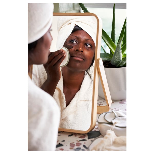 Person in white robe uses reusable pads to remove face mask, holding mirror with aloe in background.