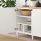 White BESTÅ sideboard with open door showing wooden bowl, books, and dish organiser on shelves. Top decorated with plants and lamp.