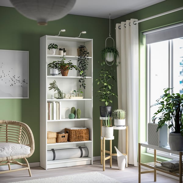 A cosy green room with a BILLY bookshelf, filled with plants and books, accompanied by a wicker chair and curtains.