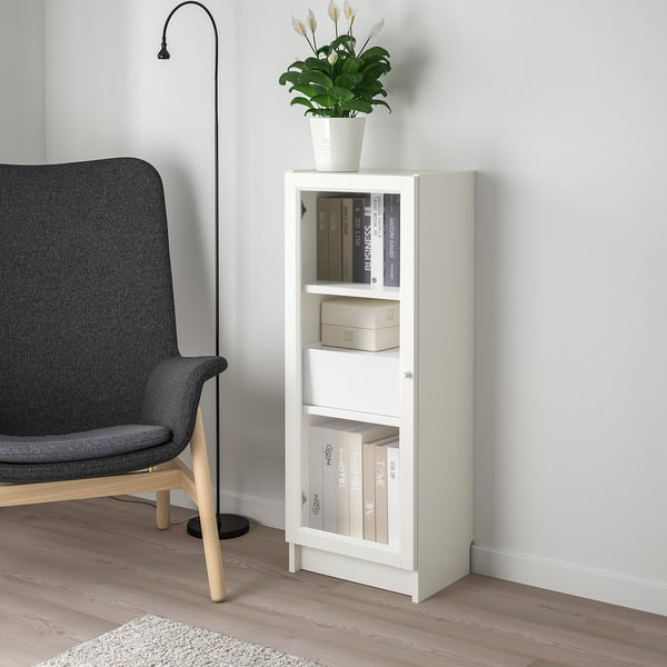 Small white BILLY bookcase, holding books & plant, stands next to grey chair. Compact, organised design.