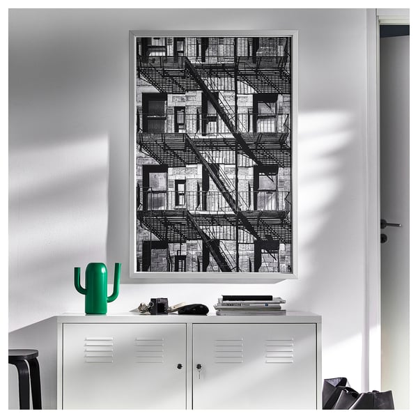 Large black-and-white framed city fire escape mounted on white wall above white locker with green cactus figurine on top.