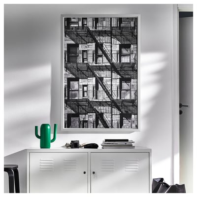 Large black-and-white framed city fire escape mounted on white wall above white locker with green cactus figurine on top.