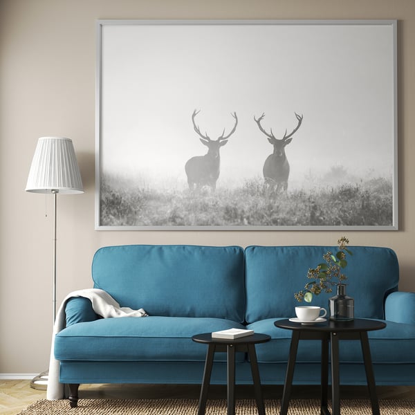 Large framed picture of two deer in foggy landscape above blue sofa. Blue sofa with black side tables, lamps, and greenery.