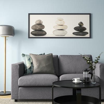 BJÖRKSTA framed art on blue wall above grey sofa. Black frame matches modern table with cup and plant. Geometric pillows, floor lamp.