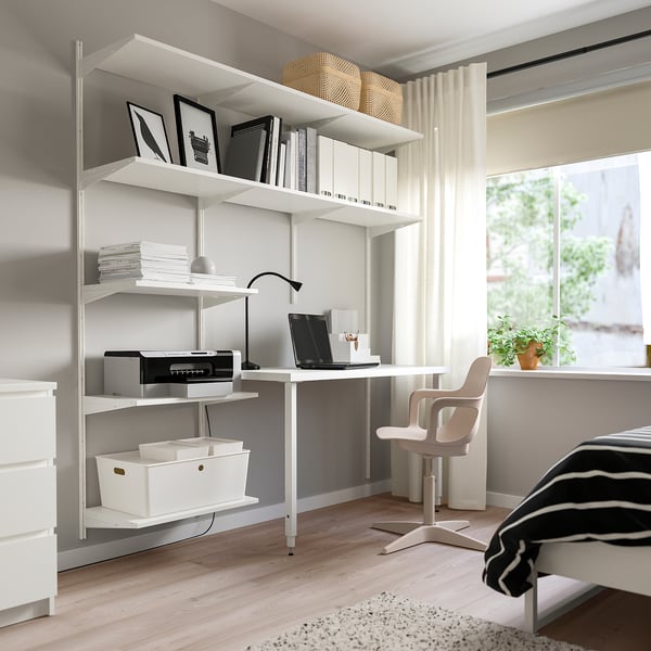 Modern home office with white BOAXEL shelves, holding books and electronics. Includes a clean desk with a chair and a bed partially visible.