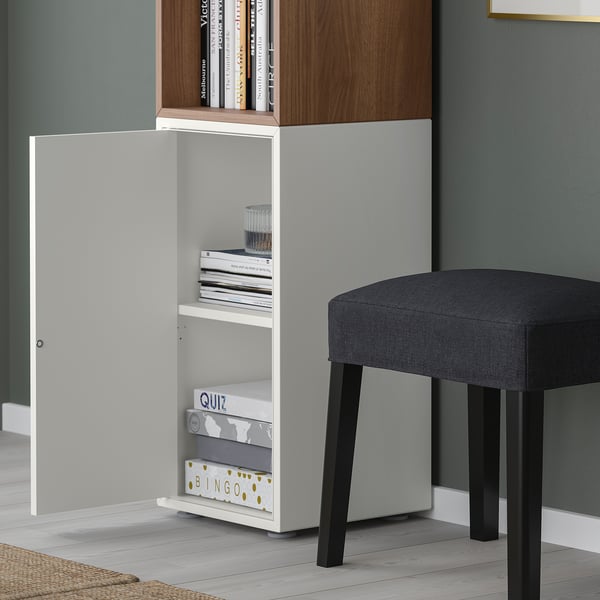 White EKET storage unit with open door, games, and books; grey upholstered stool beside it.