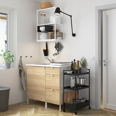 Small, multipurpose kitchen setup. Wooden unit with sink and cabinet. Open shelves with utensils. Black rolling cart with supplies.