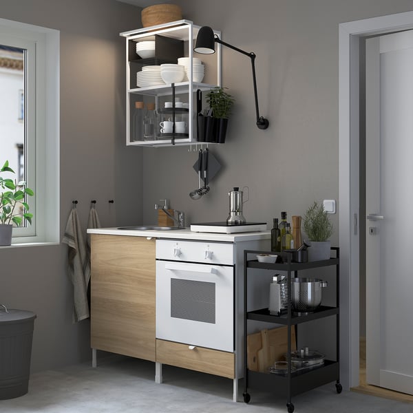 Modern compact kitchen: white oven, wood-panel base, open shelves, black cart with bottles and plants.