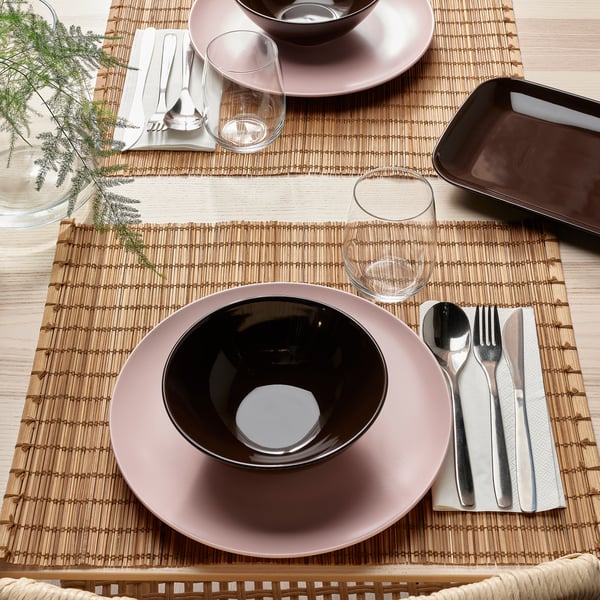 Table set with FÄRGKLAR dishes: pink plates, black bowls, silverware, glass. Natural placemats.