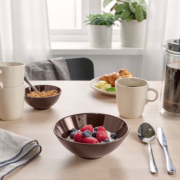 Breakfast table setup: bowl of yoghurt with berries, coffee mug, cereal bowl, croissant, cutlery, and plant.