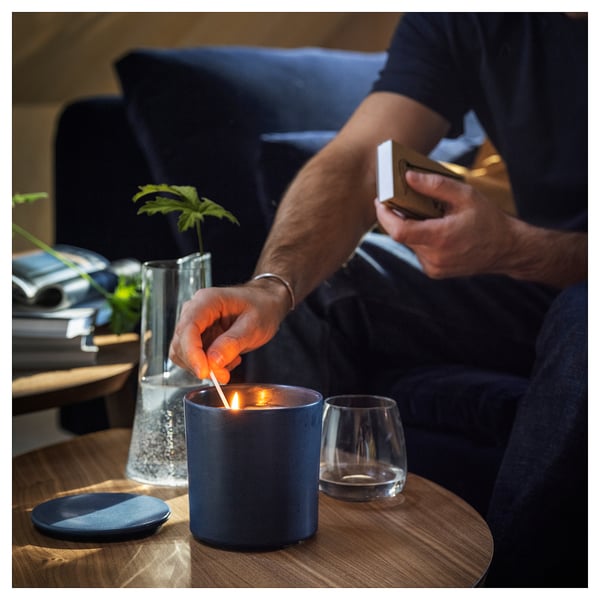 Person lights blue scented candle next to clear glass with plant, dark blue plate, and empty glass on wooden table.