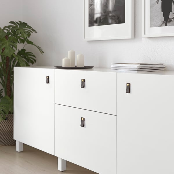 Modern white GALTHULT cabinet with black leather handles, sleek & minimalist design, fits neatly against wall with framed art & large leafy plant.