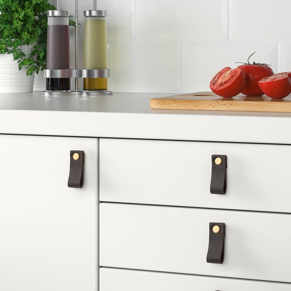 Modern kitchen with GALTHULT handles on drawers, featuring black leather grips and brass accents, with fresh tomatoes and herbs on the countertop.