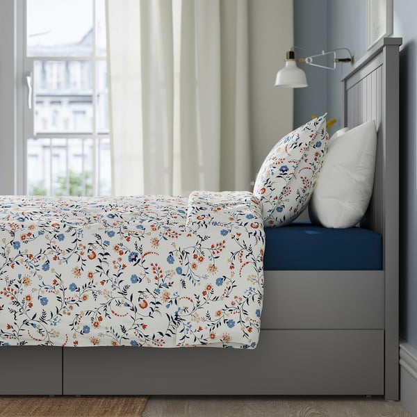 Bed with floral sheet, 2 drawers, headboard, wall lamp nearby.