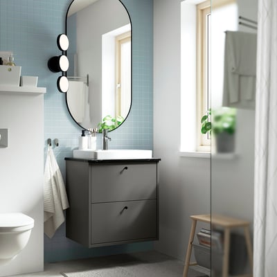 Modern bathroom with grey vanity, semi-recessed sink, oval mirror, lights, towel, & stool with books.