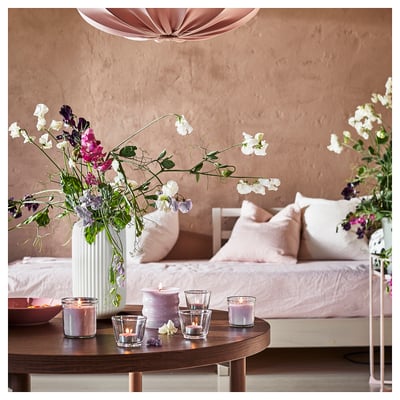 Bedroom with pink pillows, lit candles, white vase with flowers on round table, beige wall, soft lighting.