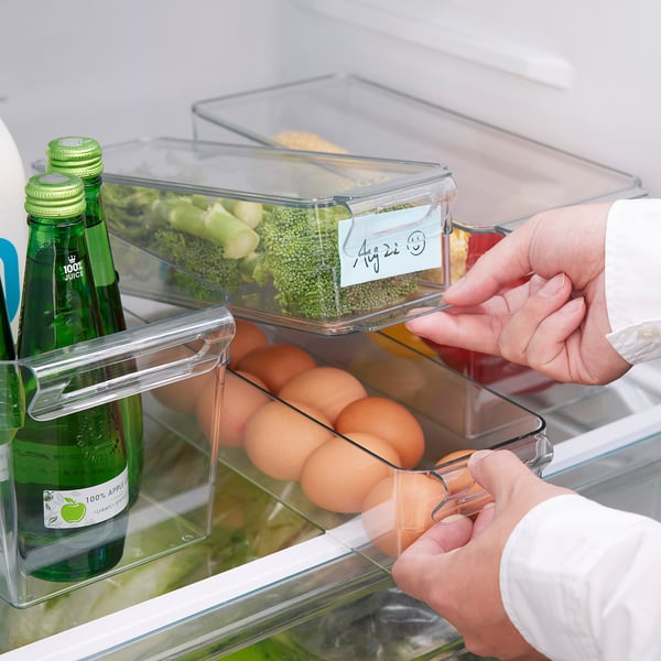 Hands place eggs in clear fridge organiser box from KLIPPKAKTUS set on a shelf, surrounded by juice bottles and broccoli.