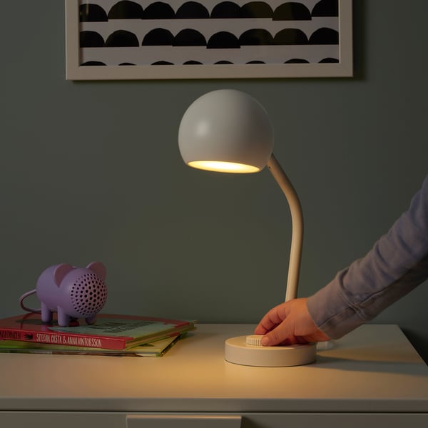 A hand adjusts a KORVSNÖRE lamp on a desk, which emits soft light. Nearby, a purple pig speaker and books are visible.