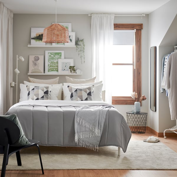 Modern bedroom with LÅSBRÄKEN grey comforter on bed, geometric pillows. Light-colored decor, wooden floor, hanging light fixture, wall art, and chair.