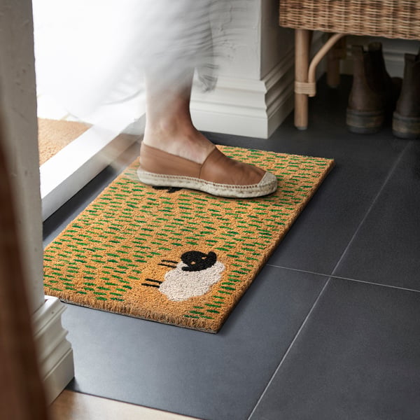 A person steps onto a colourful door mat with a sheep design, welcoming and practical for indoor use.