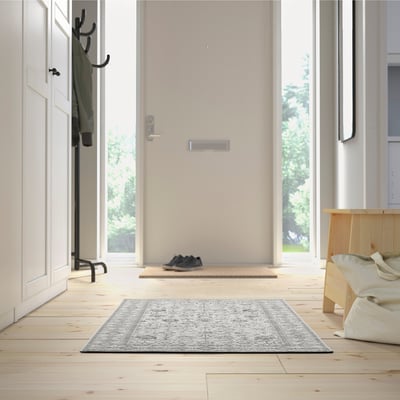 Minimalist entryway with light wood floors, grey rug, green jacket on coat rack, shoes by door, bench with beige cloth, large windows.