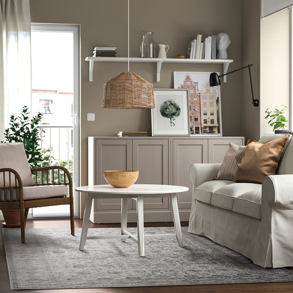 Modern living room with beige walls, brown armchair, grey sofa, wooden table, and large door to greenery. Decor includes wicker lamp and grey rug.