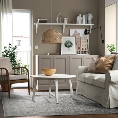 Modern living room with beige walls, brown armchair, grey sofa, wooden table, and large door to greenery. Decor includes wicker lamp and grey rug.