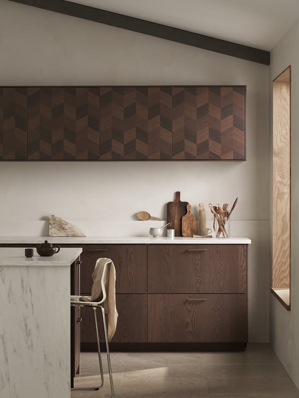 Modern kitchen with dark wood-patterned cabinets, marble countertop, wooden stool, and utensils. Herringbone design panels on upper cabinets.