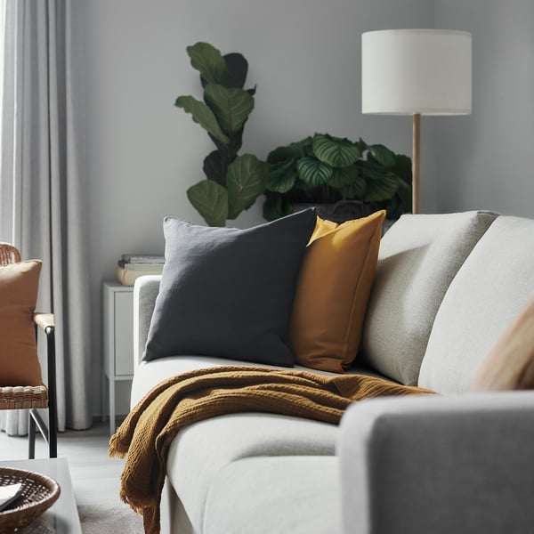 Modern living room with white couch, grey, and mustard yellow pillows, a cosy brown blanket, and a large green plant beside a lamp.