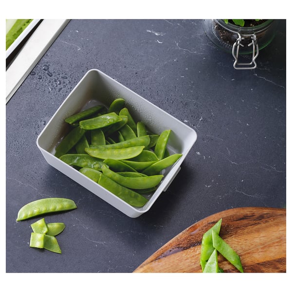 A rectangular white bowl filled with bright green snap peas on a dark grey countertop, next to a wooden cutting board with additional snap peas.