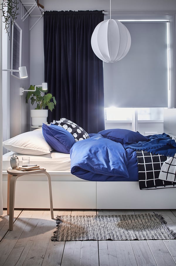 Blue velvet curtains & bedding in cosy bedroom, unmade bed with throw, plant on bedside table.