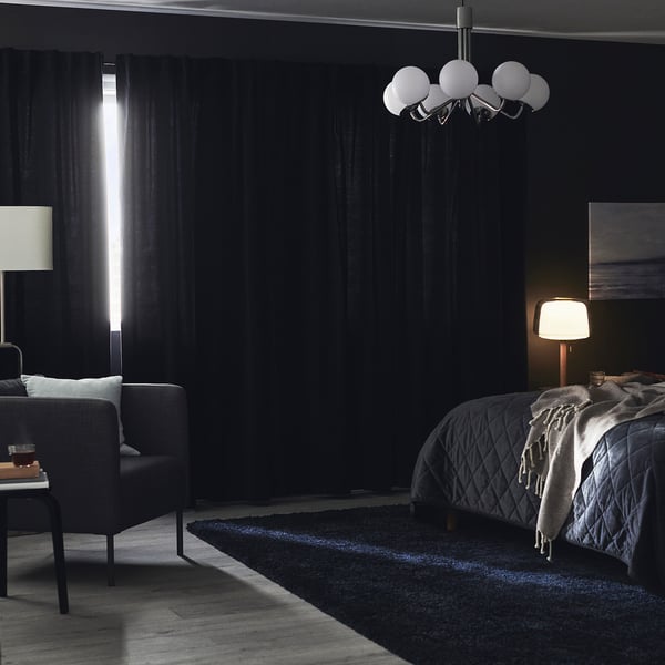 Dark bedroom with thick, blue velvet curtains blocking light, a lit table lamp, and a cosy bed with a grey blanket.