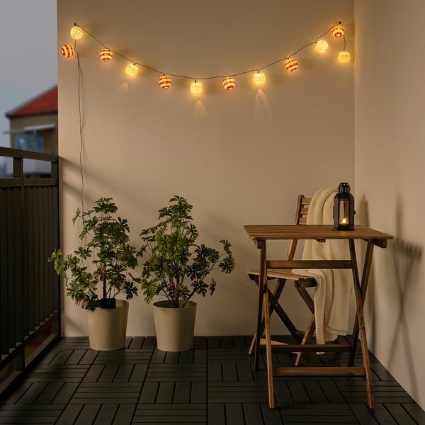 Balcony with SOLVINDEN lights, table, plants. Orange horizontal lights above table with blanket and lantern between plants.
