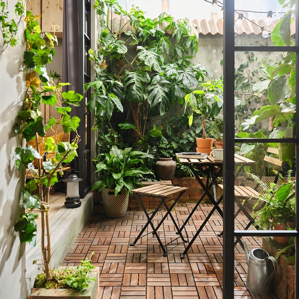 Balcony with wooden floor and furniture. TÄRNÖ stool surrounded by potted plants. Compact black stool with wooden seat and metal legs.