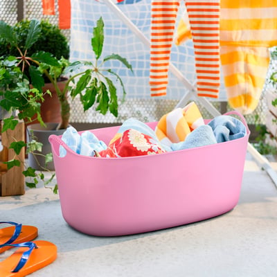 Bright pink TORKIS laundry basket with colourful towels, on concrete near flip flops.